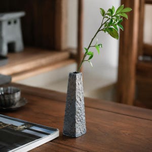 May include: A tall, slender, square-based vase with a dark, textured, stone-like finish. The vase has a tapered shape and holds a single thin green branch with small leaves extending from the top opening, creating a simple, vertical display.
