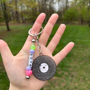 May include: A keychain featuring a miniature vinyl record charm with a silver key ring. The keychain includes a beaded strand with the word "SOUR" spelled out in white letter beads, along with pink and purple beads.