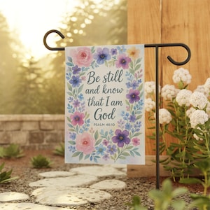 Könnte beinhalten: Eine Gartenflagge mit dem Text "Be still and know that I am God", umgeben von Aquarellblumen in Rosa, Lila und Blau. Die Flagge wird auf einem schwarzen Metallständer in einem Garten präsentiert.
