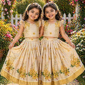 May include: Two matching two-piece outfits consisting of a sleeveless crop top with ruffled shoulders and a pleated floor-length skirt. The garments are beige with a yellow floral print and a wide gold border along the hem of the skirts.