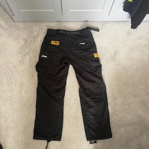 May include: Black cargo trousers with multiple pockets, a belt, and drawstrings at the ankles. The trousers have yellow and white patches with text. The trousers are laid flat on a light grey carpet.