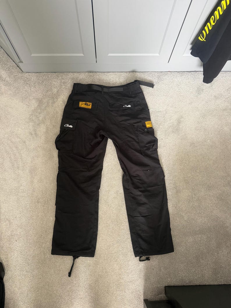 May include: Black cargo trousers with multiple pockets, a belt, and drawstrings at the ankles. The trousers have yellow and white patches with text. The trousers are laid flat on a light grey carpet.