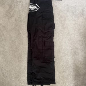 May include: Black cargo trousers with multiple pockets and a belt. A white and black graphic is on the left side of the trousers. The trousers are laid flat on a light grey surface.