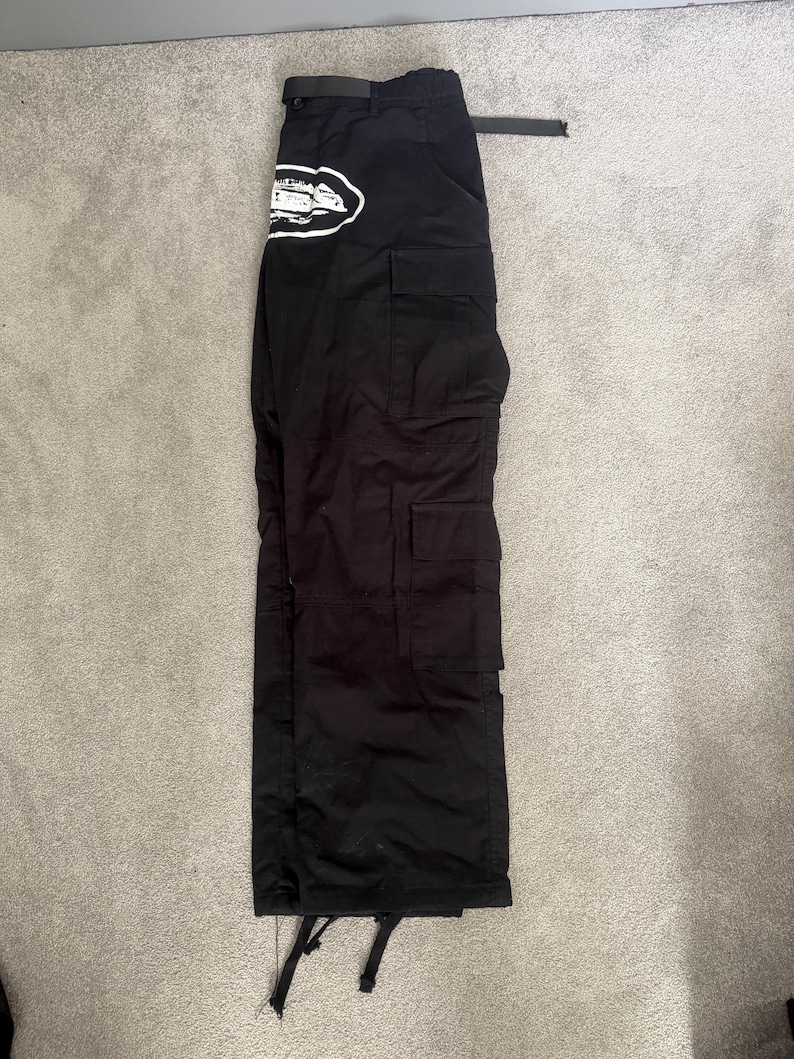 May include: Black cargo trousers with multiple pockets and a belt. A white and black graphic is on the left side of the trousers. The trousers are laid flat on a light grey surface.