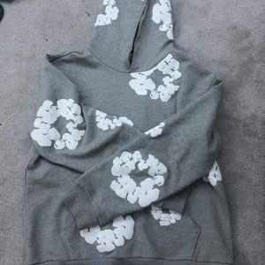 May include: A gray hoodie with white floral designs on the hood, sleeves, and chest. The hoodie has a kangaroo pocket and a drawstring hood. The fabric appears to be a soft, comfortable material.