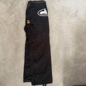 May include: Black cargo trousers with multiple pockets and a belt. A white graphic of a boat is on the upper left pocket. A yellow and black label is on the left pocket. The trousers are laid flat on a grey surface.