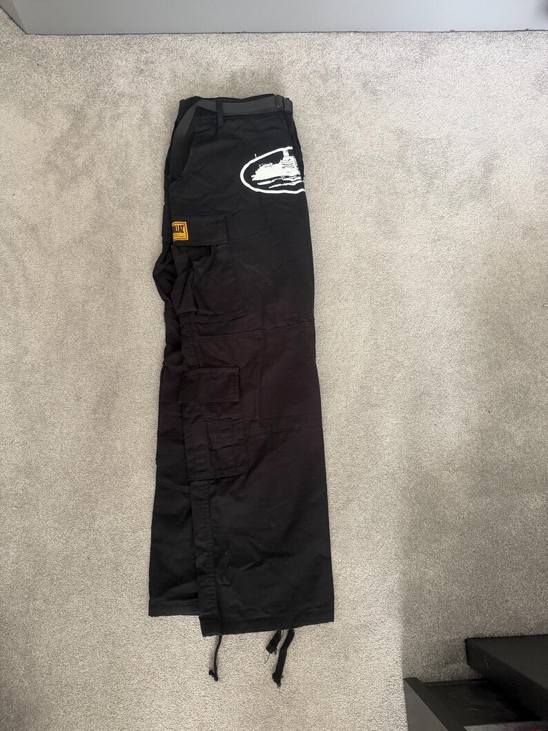 May include: Black cargo trousers with multiple pockets and a belt. A white graphic of a boat is on the upper left pocket. A yellow and black label is on the left pocket. The trousers are laid flat on a grey surface.