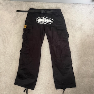 May include: Black cargo trousers with multiple pockets and a black belt. A white and black graphic is on the front. A small yellow and black patch is on the left pocket.