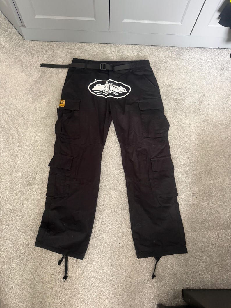 May include: Black cargo trousers with multiple pockets and a black belt. A white and black graphic is on the front. A small yellow and black patch is on the left pocket.