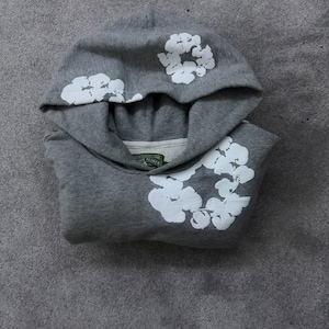 May include: A gray hoodie with white floral designs on the hood and front. The hoodie is folded and lies on a gray surface. The floral designs are clustered and stylized.