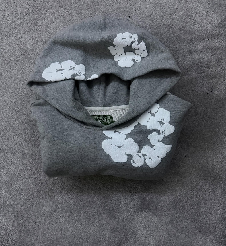 May include: A gray hoodie with white floral designs on the hood and front. The hoodie is folded and lies on a gray surface. The floral designs are clustered and stylized.