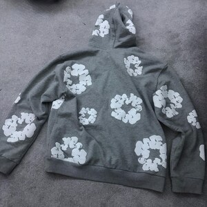 May include: A gray hoodie with a white floral pattern. The hoodie features a drawstring hood, long sleeves, and a kangaroo pocket. The white floral design is printed across the back and sleeves of the hoodie.