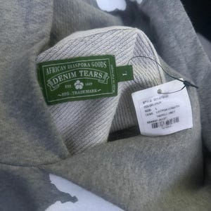 May include: A gray hoodie with white floral designs. The hoodie features a green label with the text "AFRICAN DIASPORA GOODS DENIM TEARS" and a tag with product information. The hoodie is a size L.