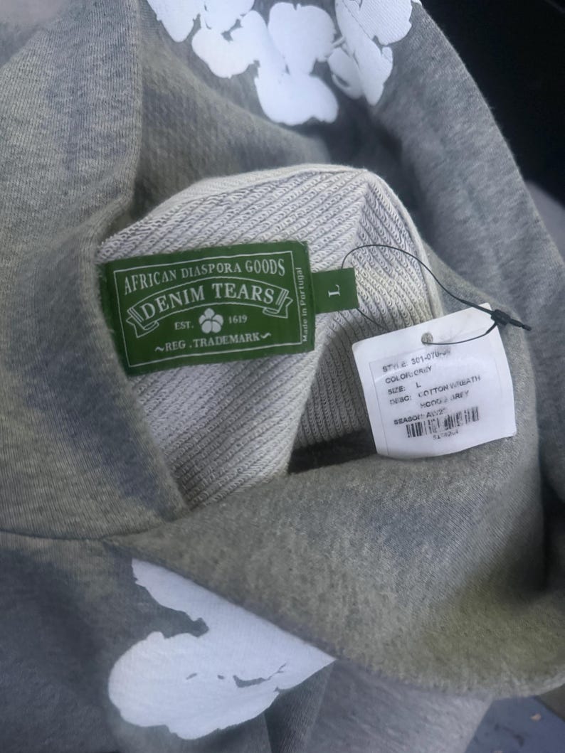 May include: A gray hoodie with white floral designs. The hoodie features a green label with the text "AFRICAN DIASPORA GOODS DENIM TEARS" and a tag with product information. The hoodie is a size L.