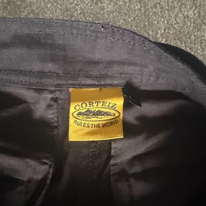 May include: Close-up of black fabric with a yellow tag. The tag features the text "CORTEIZ" above a mountain range graphic and the words "RULES THE WORID" below. The fabric appears to be part of a garment.