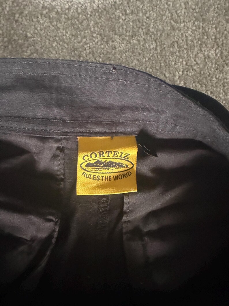 May include: Close-up of black fabric with a yellow tag. The tag features the text "CORTEIZ" above a mountain range graphic and the words "RULES THE WORID" below. The fabric appears to be part of a garment.