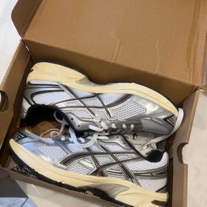May include: A pair of white and silver athletic sneakers with mesh panels and cream-colored midsoles, resting inside a brown cardboard shoe box. The sneakers feature dark grey accents, white laces, and a structured design with visible side branding.