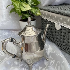 Op de afbeelding: Een gepolijste zilveren theepot met een gebogen handvat met zwarte accenten, een decoratieve knop op het deksel en drie versierde voetjes. Het lichaam van de theepot heeft een glad, reflecterend oppervlak met gedetailleerde afwerkingen.