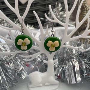 May include: A pair of dangle earrings featuring circular green discs with a three-petaled cream-colored flower design and a small square lime green center stone. The earrings hang from silver-toned metal hooks.