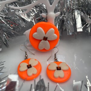 May include: A jewelry set consisting of a pendant and two matching earrings. Each piece features a bright neon orange circular base with a three-petaled cream-colored flower design in the center, accented by a small square brown stone.