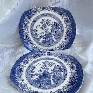 May include: Two blue and white Willow pattern plates. The plates have a rounded square shape with a detailed border. The central design features a scenic landscape with a bridge, pagoda, and birds. The plates are set against a white background.