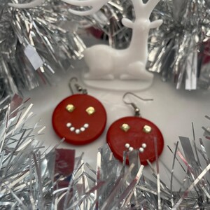 Peut inclure: Une paire de boucles d'oreilles rondes et rouges arborant un motif de visage souriant. Chaque boucle possède deux petits clous dorés en guise d'yeux et une rangée incurvée de points blancs formant une bouche, avec des crochets en métal argenté.