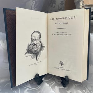 May include: An open hardback book titled The Moonstone by Wilkie Collins. The left page features a black and white ink portrait of the author, William Wilkie Collins. The right page displays the title, author, introduction credits, and publisher.