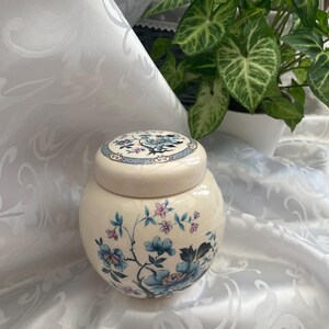May include: A cream-colored ceramic jar with a matching lid, featuring a delicate floral pattern in shades of blue, pink, and dark green. The rounded vessel displays intricate botanical illustrations of blooming flowers and thin branches.