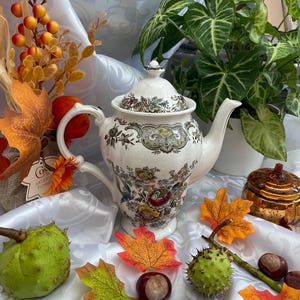 May include: A white ceramic teapot featuring a detailed floral and botanical pattern in shades of green, brown, and yellow. The teapot has a matching lid with a small knob, a curved handle, and a spout, surrounded by autumn leaves and chestnuts.