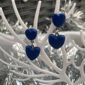 May include: A pair of drop earrings featuring dark blue heart-shaped charms with a subtle glitter finish. One earring consists of two stacked hearts connected by a small metal jump ring, while the other features a single heart charm.