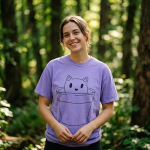 Könnte beinhalten: Ein lilafarbenes T-Shirt mit einer schwarzen Strichzeichnung einer Katze in einem Blumentopf. Die Katze hat ein einfaches, lächelndes Gesicht. Das kurzärmelige Shirt ist aus einem weichen Material.