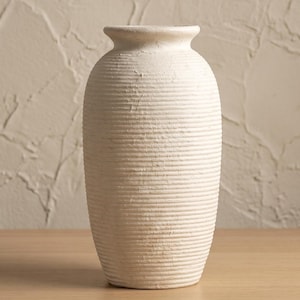 May include: A cream-colored, textured vase with a ribbed design. The vase has a rounded body and a narrow neck. It is sitting on a light-colored wooden surface. The background is a textured wall.