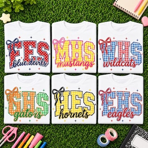 May include: Six white t-shirts featuring school acronyms in block letters with gingham patterns and bows. The designs include FES blue devils, MHS mustangs, WHS wildcats, GHS gators, HES hornets, and EHS eagles, all accented with small colourful stars.
