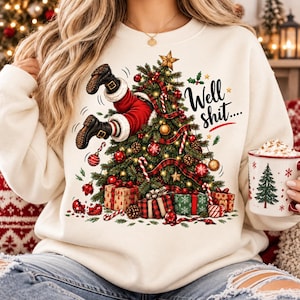 May include: A cream-colored sweatshirt featuring a graphic of a Christmas tree with Santa's legs sticking out of the top. The design includes red and green ornaments, wrapped gifts, and the text Well shit in black script with a red underline.