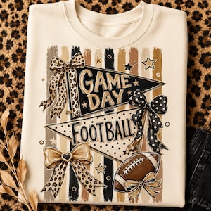 May include: A cream-coloured crewneck sweatshirt featuring a graphic print of two pennants with the text GAME DAY and FOOTBALL. The design includes leopard print bows, a black polka dot bow, and a brown football, all set against vertical stripes.
