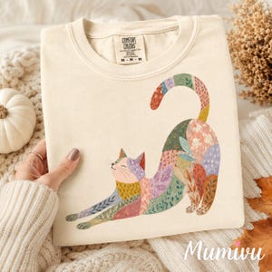 May include: A cream-coloured crewneck t-shirt featuring a graphic of a stretching cat. The cat is decorated with various colourful floral and leaf patterns in shades of pink, green, orange, and purple. The inner neck label reads COMFORT COLORS.