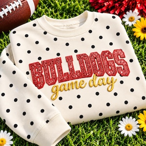 Op de afbeelding: Een crèmekleurige sweater met ronde hals en een zwart stippenpatroon. Op de voorzijde staat het woord BULLDOGS in rode glitterende blokletters met een dikke rode rand, en de woorden game day in gele cursieve letters eronder.
