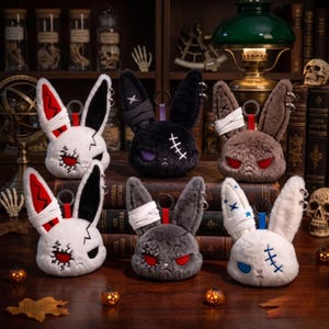 May include: Six plush rabbit head keychains with varied designs, including white, black, gray, and brown fur. Each features stitched details, metal ring attachments, and unique eye colors like red, purple, and blue, arranged on stacked books.