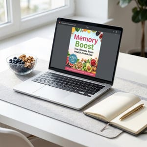 Könnte beinhalten: Ein Laptop zeigt das Cover eines Buches mit dem Titel "Memory Boost: The Ultimate Brain Health Diet Guide". Das Cover zeigt farbenfrohe Illustrationen von Früchten, Nüssen und Gemüse. Eine Schüssel mit Blaubeeren und Walnüssen steht daneben, zusammen mit einem Notizbuch und einem Stift.