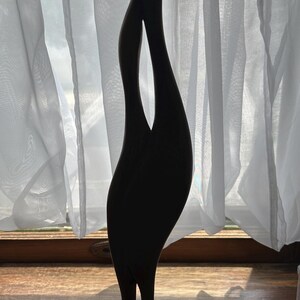 May include: A dark wooden sculpture of two intertwined figures, standing on a light-colored, rounded base. The sculpture has a smooth, flowing design with elongated, upward-reaching forms. The background features a window with sheer white curtains.