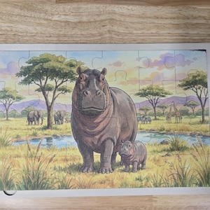 May include: A rectangular jigsaw puzzle featuring a colorful illustration of a large adult hippopotamus and a small calf standing in a grassy savanna landscape with a river, acacia trees, elephants, giraffes, and zebras in the background.
