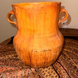 May include: A rustic, orange-toned ceramic vase with two curved handles. The vase has a wide, rounded body and a slightly flared rim. The surface shows variations in color and texture, suggesting an aged appearance. The vase is displayed on a patterned fabric.