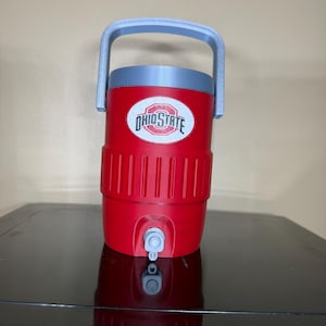 May include: A red water cooler with a gray handle and lid. The cooler has the words "OHIO STATE" in a white oval. A silver spigot is at the bottom. The cooler is on a black surface.