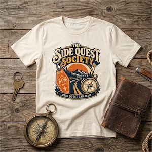 Peut inclure: Un t-shirt à manches courtes couleur crème avec une impression graphique portant le texte THE SIDE QUEST SOCIETY et MAIN QUEST CAN WAIT. Le motif inclut un paysage de montagne, un porte-clés de motel et une boussole.