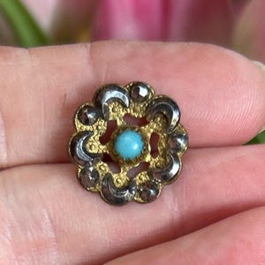 Op de afbeelding: Een kleine, antieke broche met een bloemmotief. De broche heeft een centrale, ronde, lichtblauwe steen omgeven door een goudkleurige metalen zetting. De buitenrand heeft een zilverkleurig metaal met decoratieve accenten.