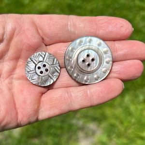May include: Two round, iridescent mother-of-pearl buttons with four central holes. The smaller button features a geometric etched pattern, while the larger button has a smooth, polished surface with a ring of small, circular indentations.