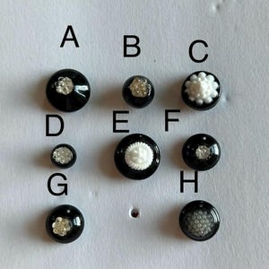 以下が含まれることがあります： AからHまでのラベルが付いた8つの丸い黒いボタンのコレクション。それぞれにユニークな白または透明のテクスチャ加工された中央のディテールが施されています。ボタンはサイズとデザインが異なり、複雑なビーズのようなパターンや、花をモチーフにした白い中心部が見られます。