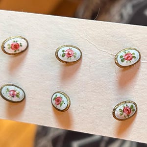 May include: Six oval-shaped buttons featuring a white enamel surface with a hand-painted red rose and green leaves motif. Each button is framed by a textured gold-tone metal border and is attached to a piece of off-white cardstock for display.