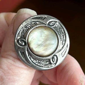 Puede incluir: Un botón redondo de plata antigua con un centro nacarado iridiscente. El botón tiene un diseño ornamentado en relieve alrededor del borde, con espirales y detalles punteados. El botón es probablemente vintage y adecuado para coser o manualidades.