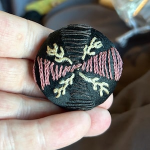 May include: A circular black fabric-covered button featuring decorative embroidery. The design includes two horizontal sections of pink thread stitching and four cream-coloured floral sprig motifs arranged in a cross pattern on the black surface.